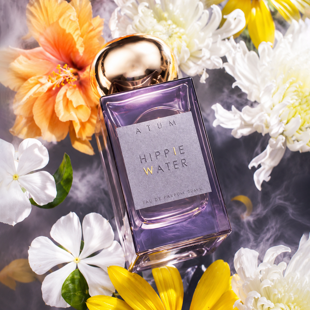 A rectangular glass perfume bottle labeled 'ATUM Hippie Water' with a metallic gold cap, surrounded by yellow and white flowers on a smoky gray background.