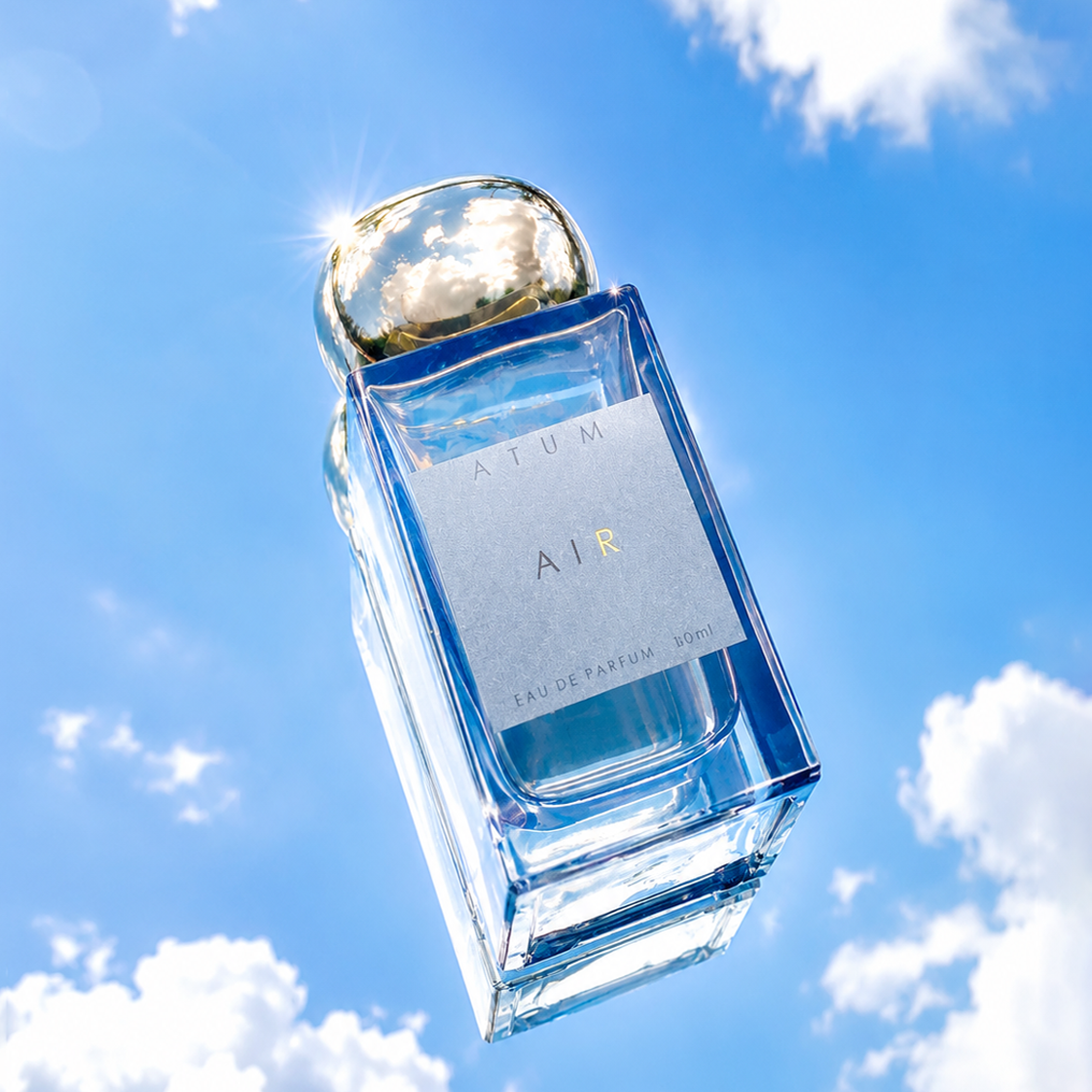 A clear glass perfume bottle labeled ATUM AIR, floating against a bright blue sky with white clouds. The bottle has a metallic gold cap and light blue liquid inside.