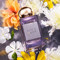A rectangular glass perfume bottle labeled 'ATUM Hippie Water' with a metallic gold cap, surrounded by yellow and white flowers on a smoky gray background.