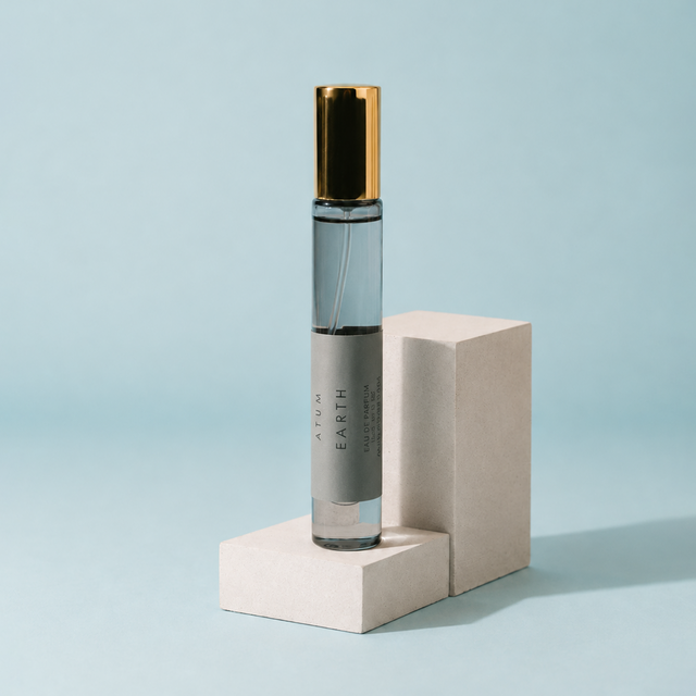 A clear glass roller bottle with a metallic gold cap and a gray label reading 'ATM EARTH', displayed on two stacked beige blocks against a light blue background.