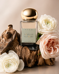 A rectangular glass perfume bottle labeled ATUM Forever Flowers, with a reflective gold cap, styled on a piece of driftwood and surrounded by white and blush pink flowers against a warm beige background.