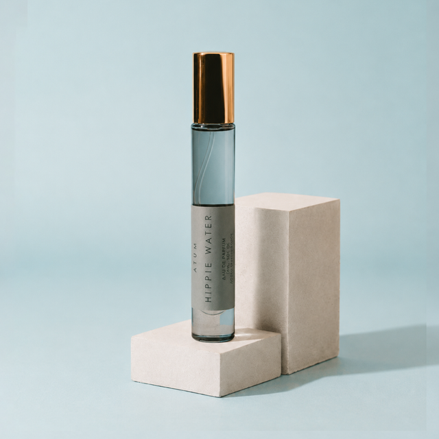 A cylindrical roll-on perfume bottle labeled 'HIPPIE WATER' by AURUM, standing on a light beige pedestal against a pale blue background.