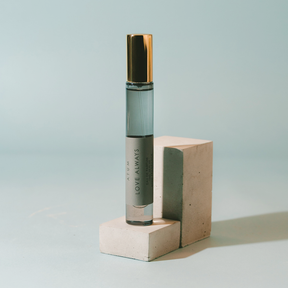 A clear glass roll-on perfume bottle with a metallic gold cap and a gray label reading 'ARUM LOVE ALWAYS' and 'PARFUM', displayed on two stacked concrete blocks against a light blue background.