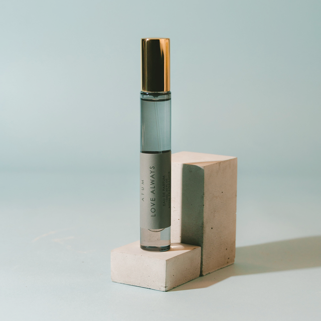 A clear glass roll-on perfume bottle with a metallic gold cap and a gray label reading 'ARUM LOVE ALWAYS' and 'PARFUM', displayed on two stacked concrete blocks against a light blue background.