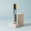 A clear cylindrical perfume or roll-on bottle with a metallic gold cap stands on a light beige geometric pedestal against a pale blue background. The bottle label reads 'WHITE SANDS' and 'ALYM'.
