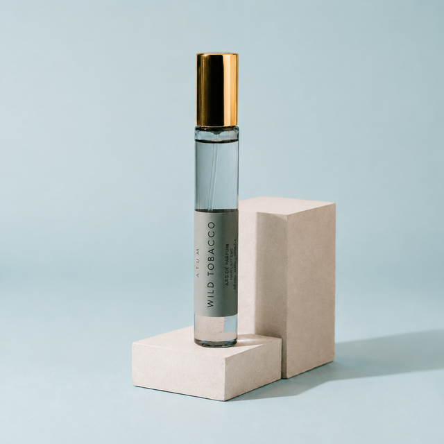 A clear glass roller bottle with a metallic gold cap and a gray label reading 'WILD TOBACCO' and 'AROMAS PARFUM', displayed on two light beige blocks against a pale blue background.