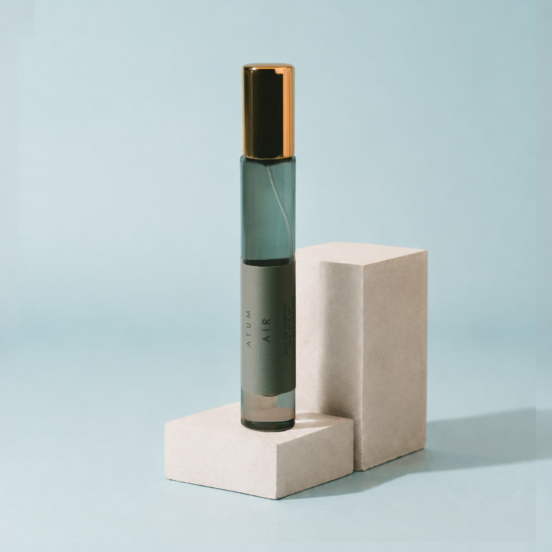 A cylindrical glass roller bottle with a dark green-gray body and a metallic gold cap, standing on a light beige geometric display block against a pale blue background. The bottle is labeled 'ATOM AIR'.