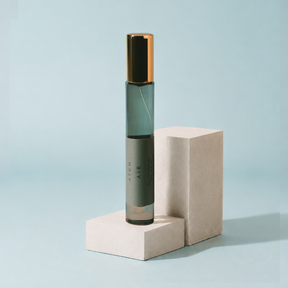 A cylindrical glass roller bottle with a dark green-gray body and a metallic gold cap, standing on a light beige geometric display block against a pale blue background. The bottle is labeled 'ATOM AIR'.