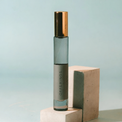 A clear glass roller bottle with a metallic gold cap and a gray label reading 'OFFERINGS', standing on a light concrete block against a pale blue background.