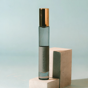 A clear glass roller bottle with a metallic gold cap and a gray label reading 'OFFERINGS', standing on a light concrete block against a pale blue background.