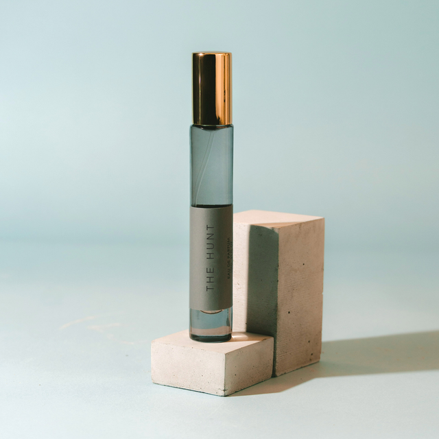 A glass rollerball perfume bottle with a metallic gold cap and a gray label reading 'THE HUNT' stands on two light beige concrete blocks against a pale blue background.
